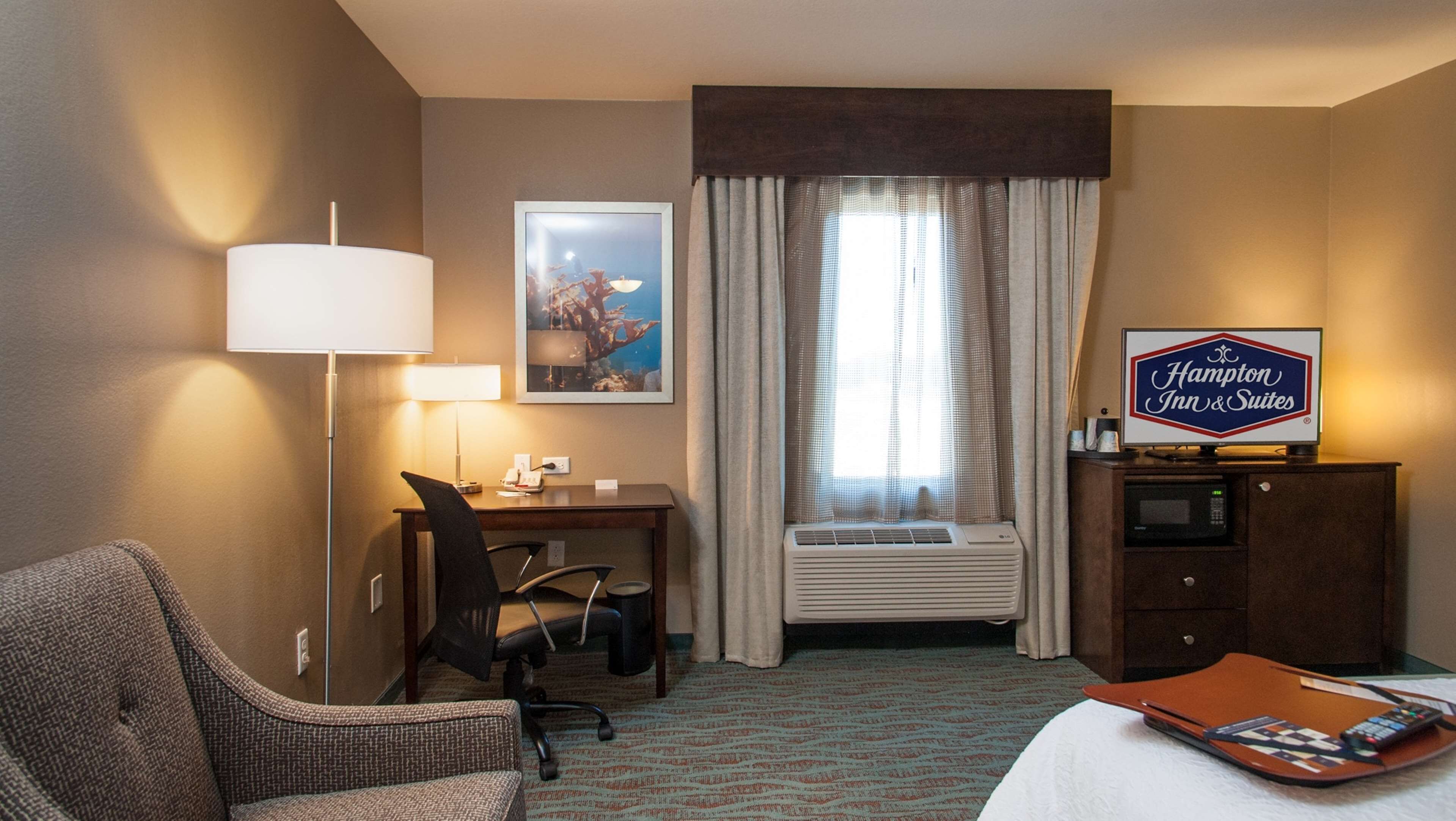 A hotel room with a desk, chair, lamp, air conditioning unit, and a sign for Hampton Inn & Suites.