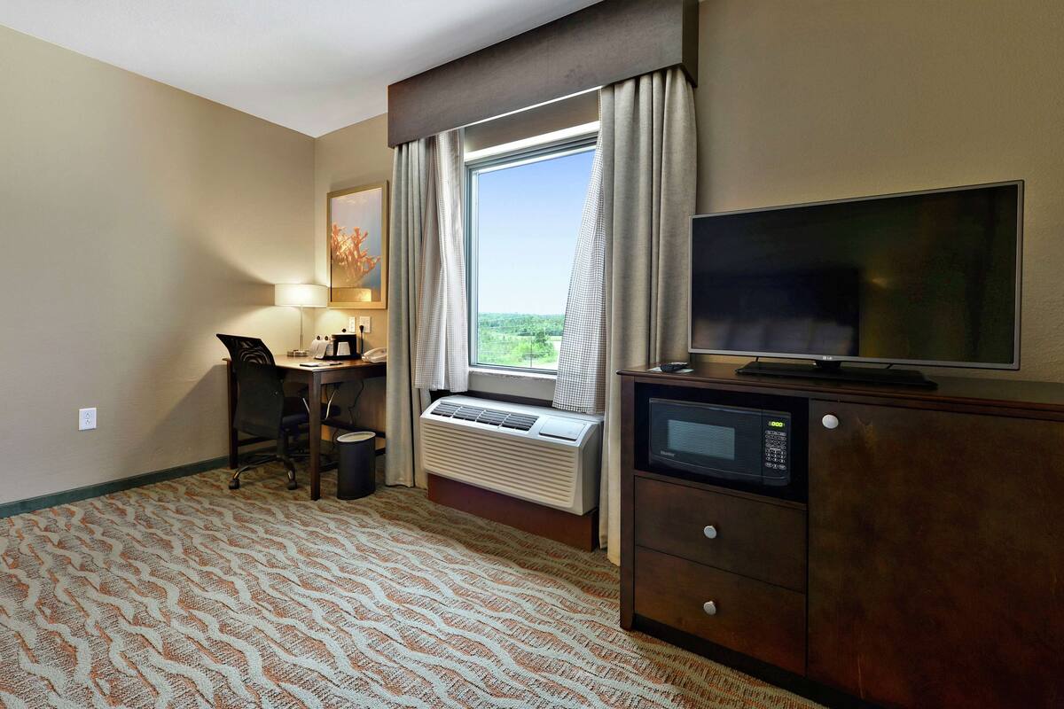 Hotelkamer met groot raam, bureau met stoel, televisie, magnetron en kast.