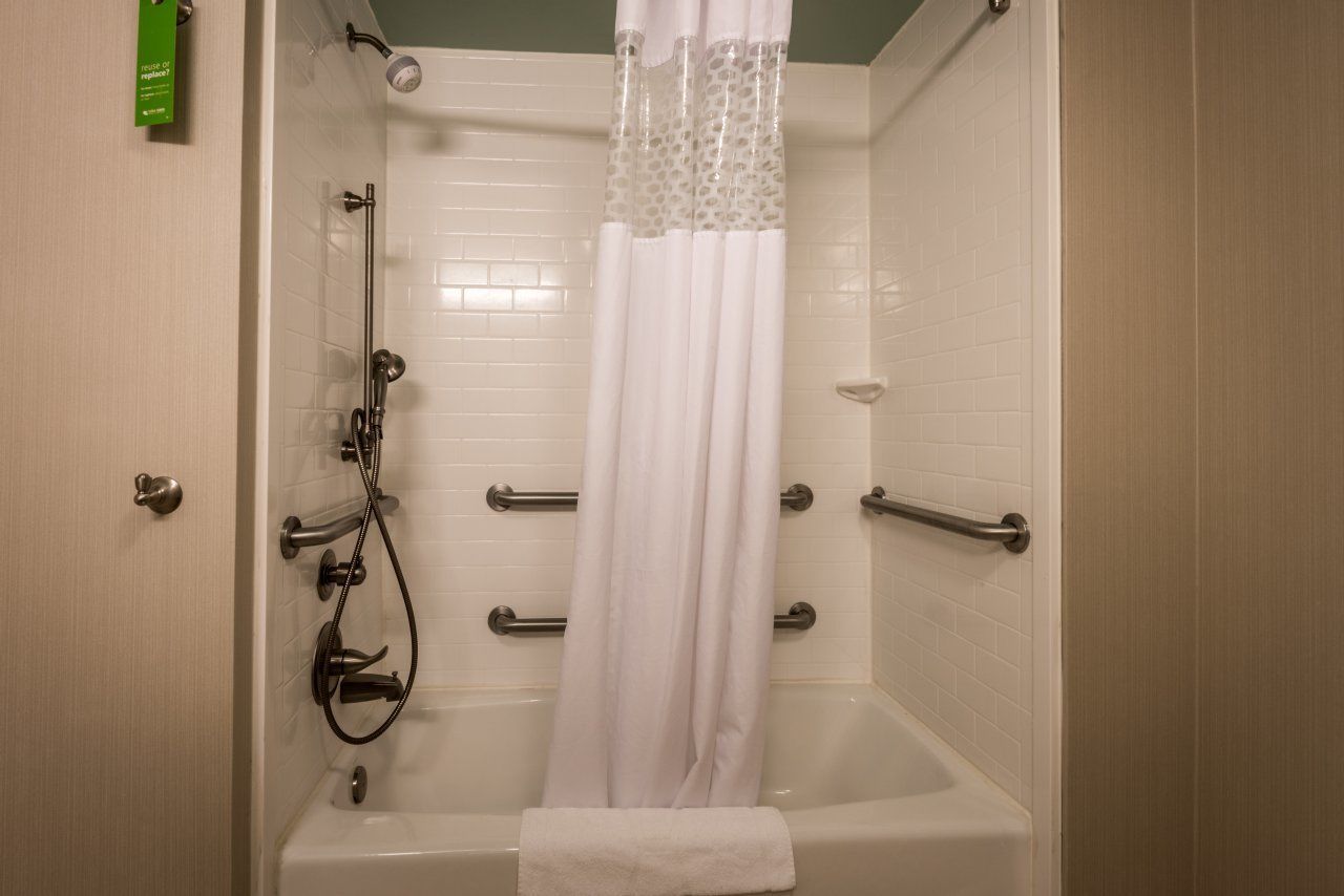 A walk-in shower with a white tiled wall, a showerhead, a handheld showerhead, a glass shower curtain, and a towel hanging on the wall.