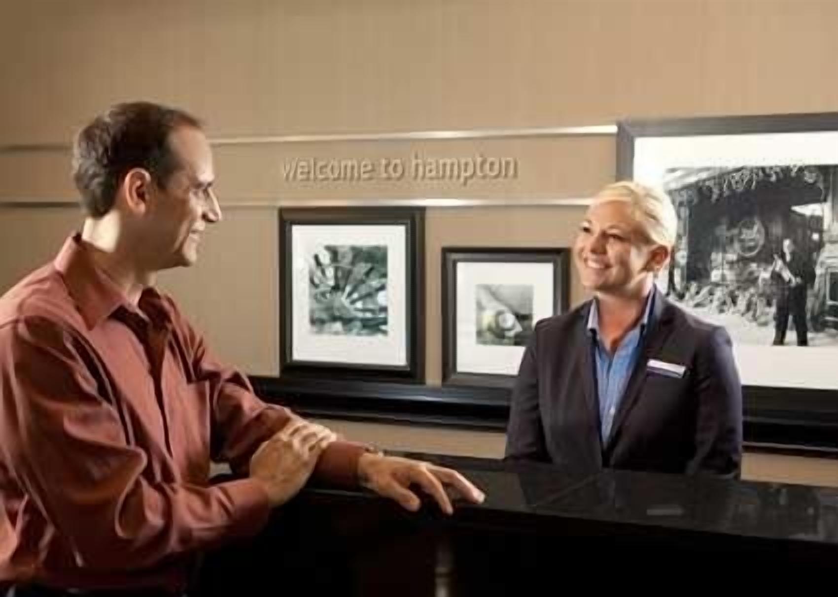 Two individuals at a reception desk with framed artwork on the wall and a 'Welcome to Hampton' sign.