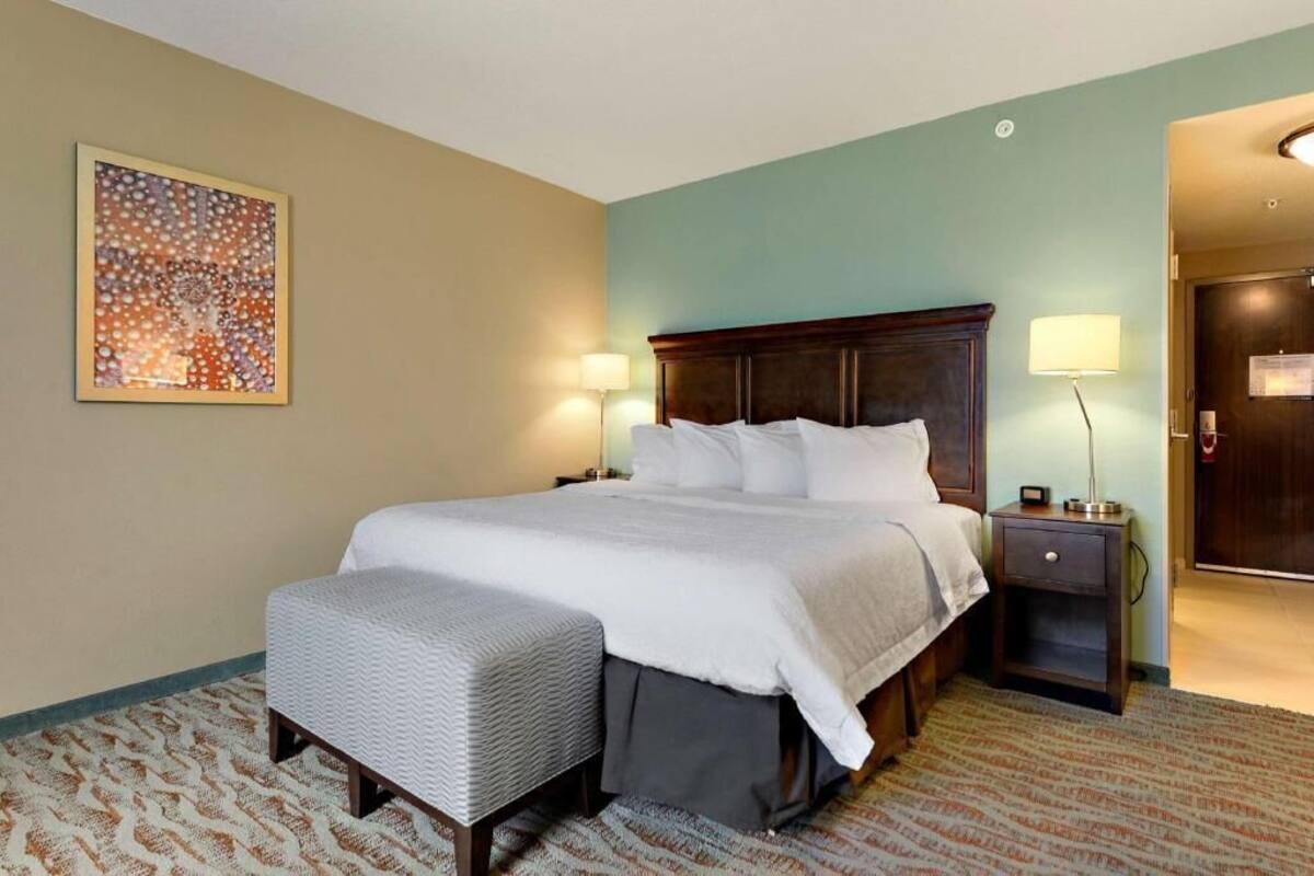 Een hotelkamer met een groot bed, nachtkastjes, een bankje, een kunstwerk aan de muur en een deur.