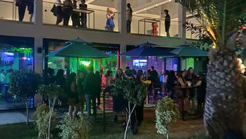 A multi-level outdoor area with people gathered around tables and chairs, surrounded by greenery and a palm tree.