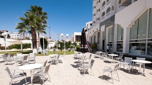 An outdoor patio area with multiple tables and chairs, palm trees, and a building with large windows. - City Business Monastir Center (Monastir)