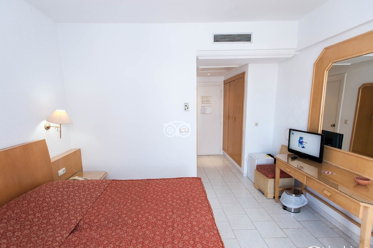 A hotel room with a bed, a desk with a computer, a mirror, and a small kitchenette area.