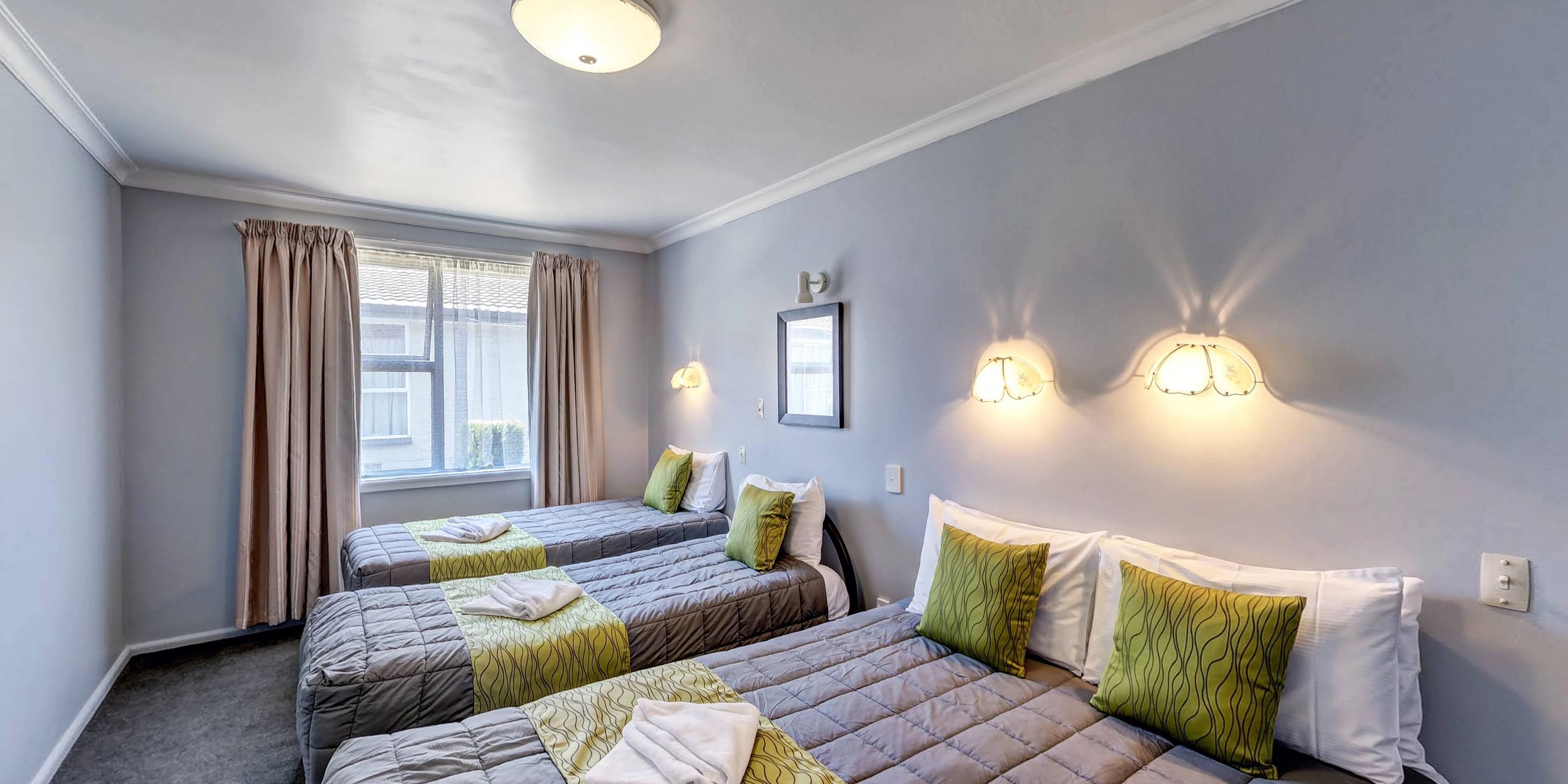 Une chambre d’hôtel avec deux lits, des rideaux beiges, une fenêtre et des lampes fixées au mur.