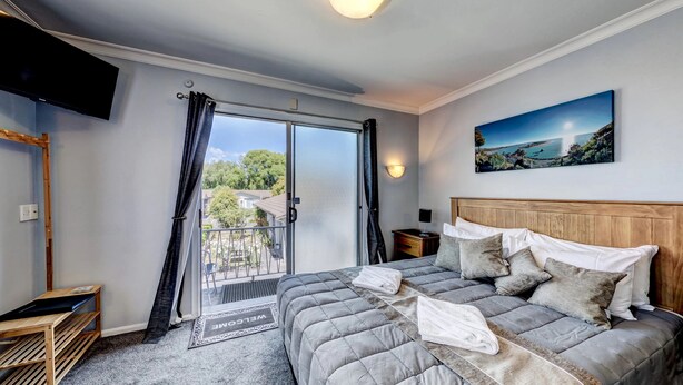 A bedroom with a large bed, a TV mounted on the wall, a balcony with a view of greenery, and a painting above the bed. - Admiral Motel (Christchurch)
