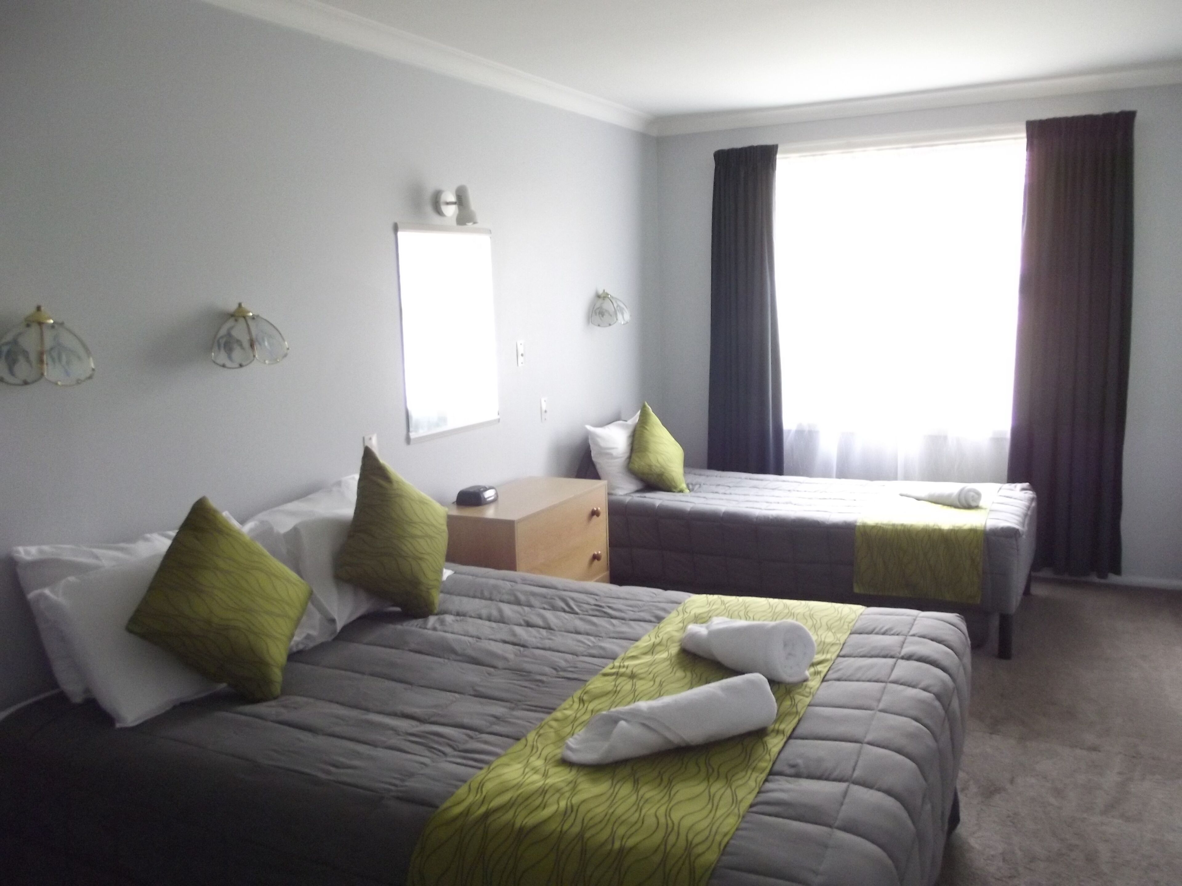 A hotel room with two beds, each with a green and white bedspread, and a large window with dark curtains.