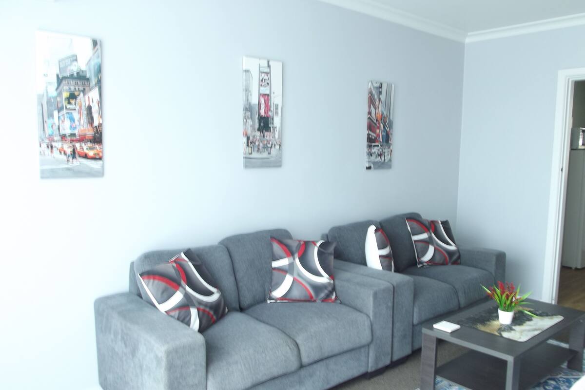 A grey sofa with red and white patterned cushions, a small table with a plant, and three framed pictures on the wall.