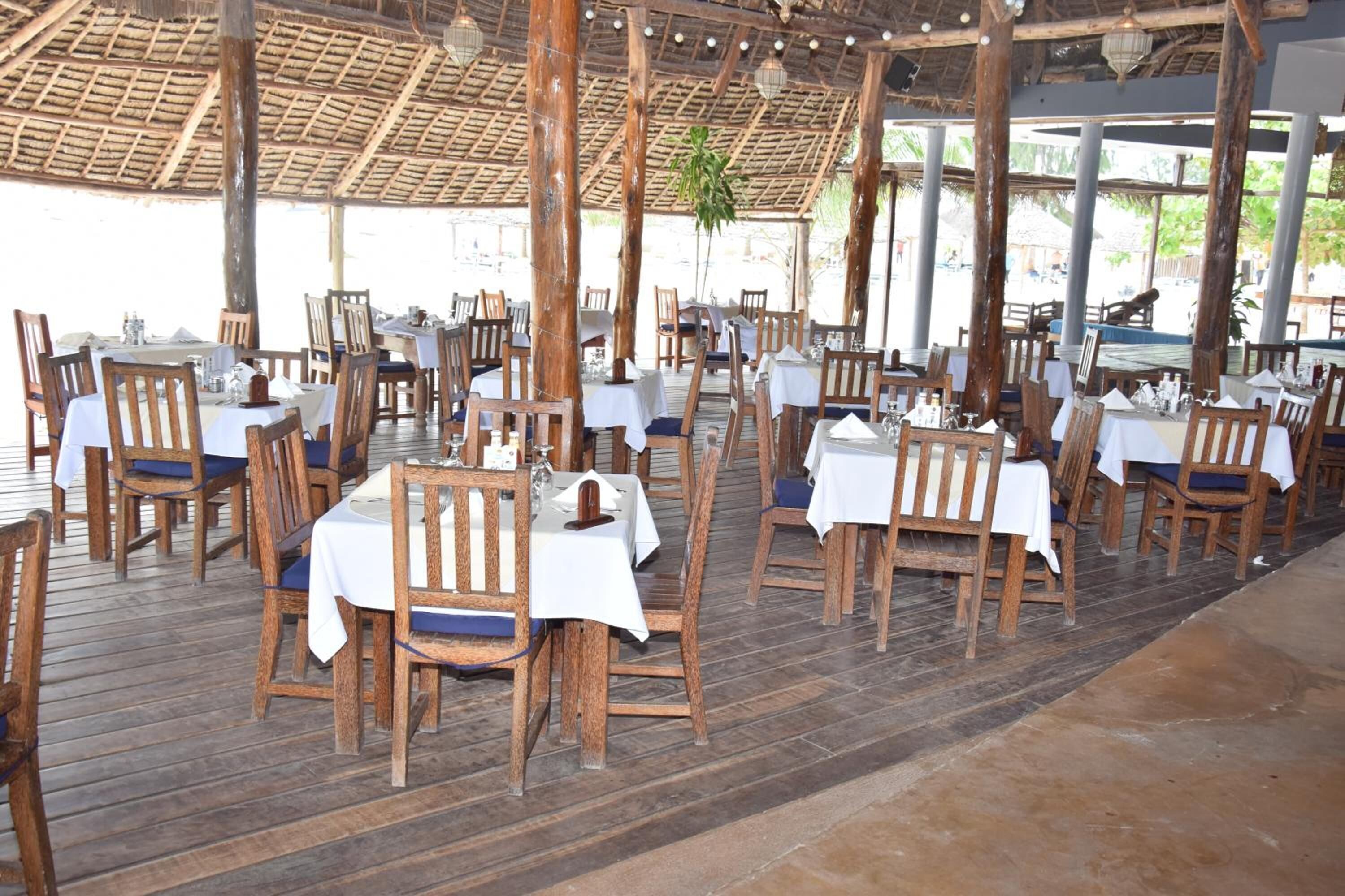 An outdoor dining area with wooden tables and chairs, covered by a thatched roof.