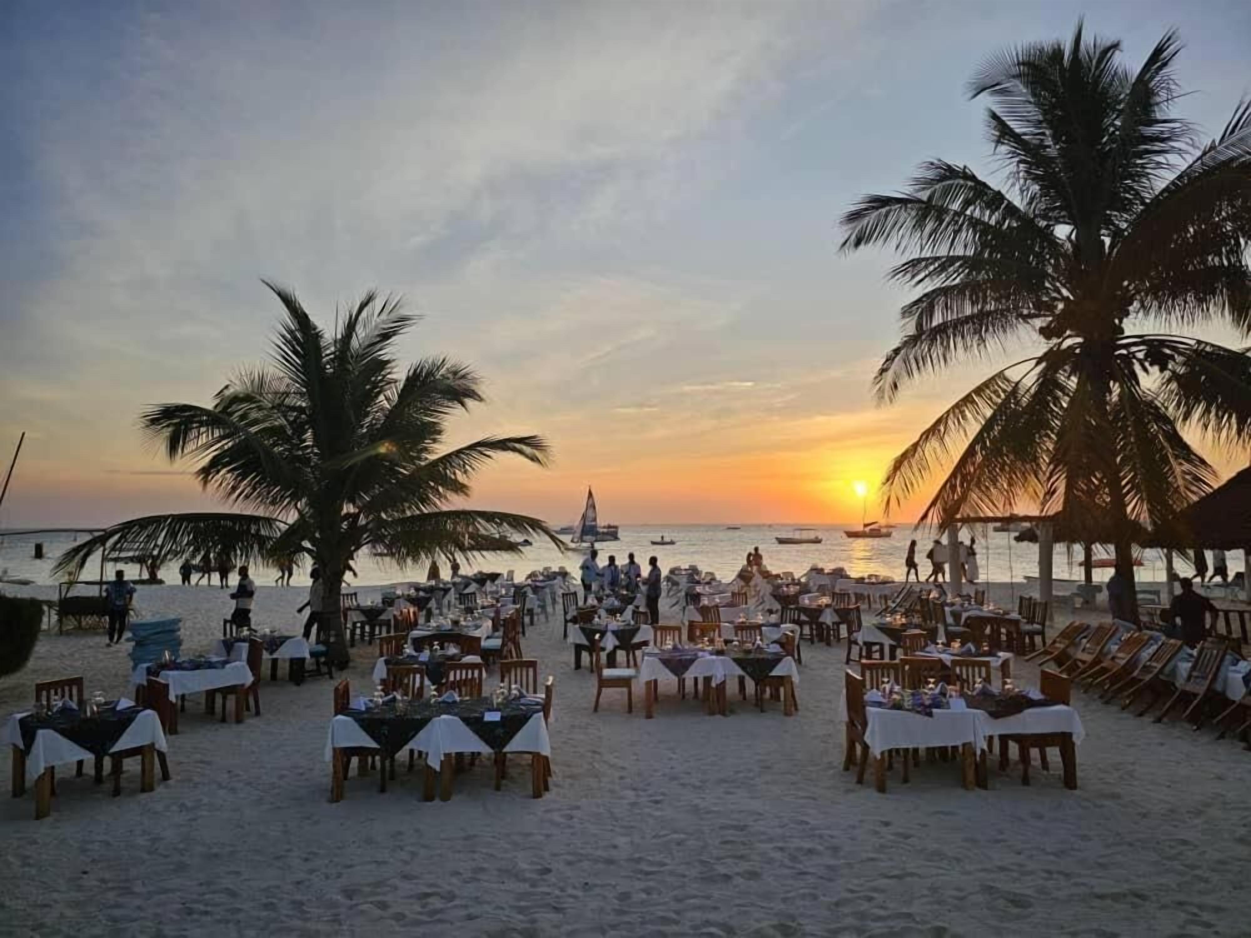 En restaurant ved stranden med borde og stole, palmer og en solnedgang i baggrunden.