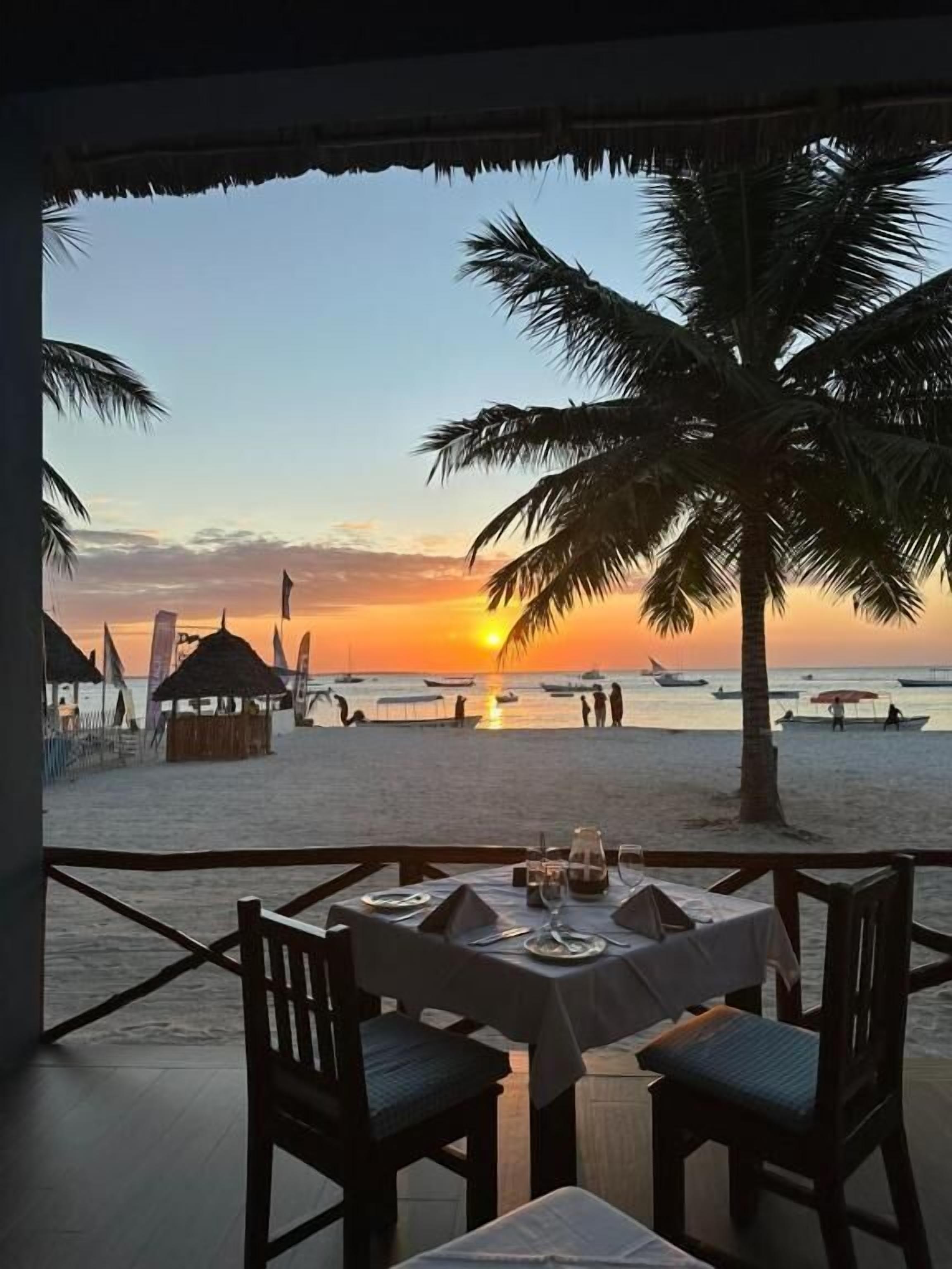 A dining area with tables and chairs, overlooking a beach with palm trees and a sunset.