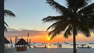 Comedor con mesas y sillas, con vista a una playa con palmeras y un atardecer.