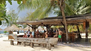 Un bar junto a la playa con bancos de madera y techo de paja, rodeado de palmeras y una playa arenosa.