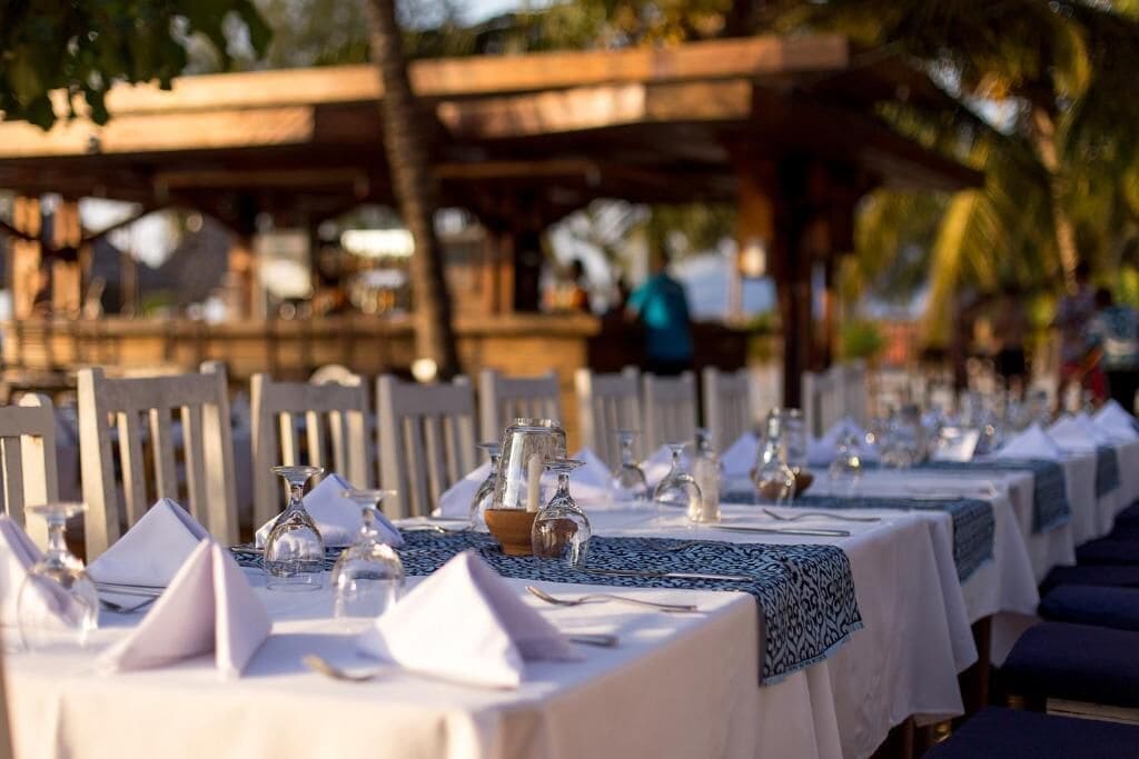 A row of tables set for dining outdoors with white tablecloths and blue patterned runners.