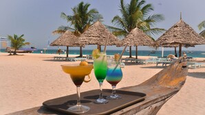 Escena de playa con un banco de madera que muestra bebidas coloridas, palmeras y un barco al fondo.