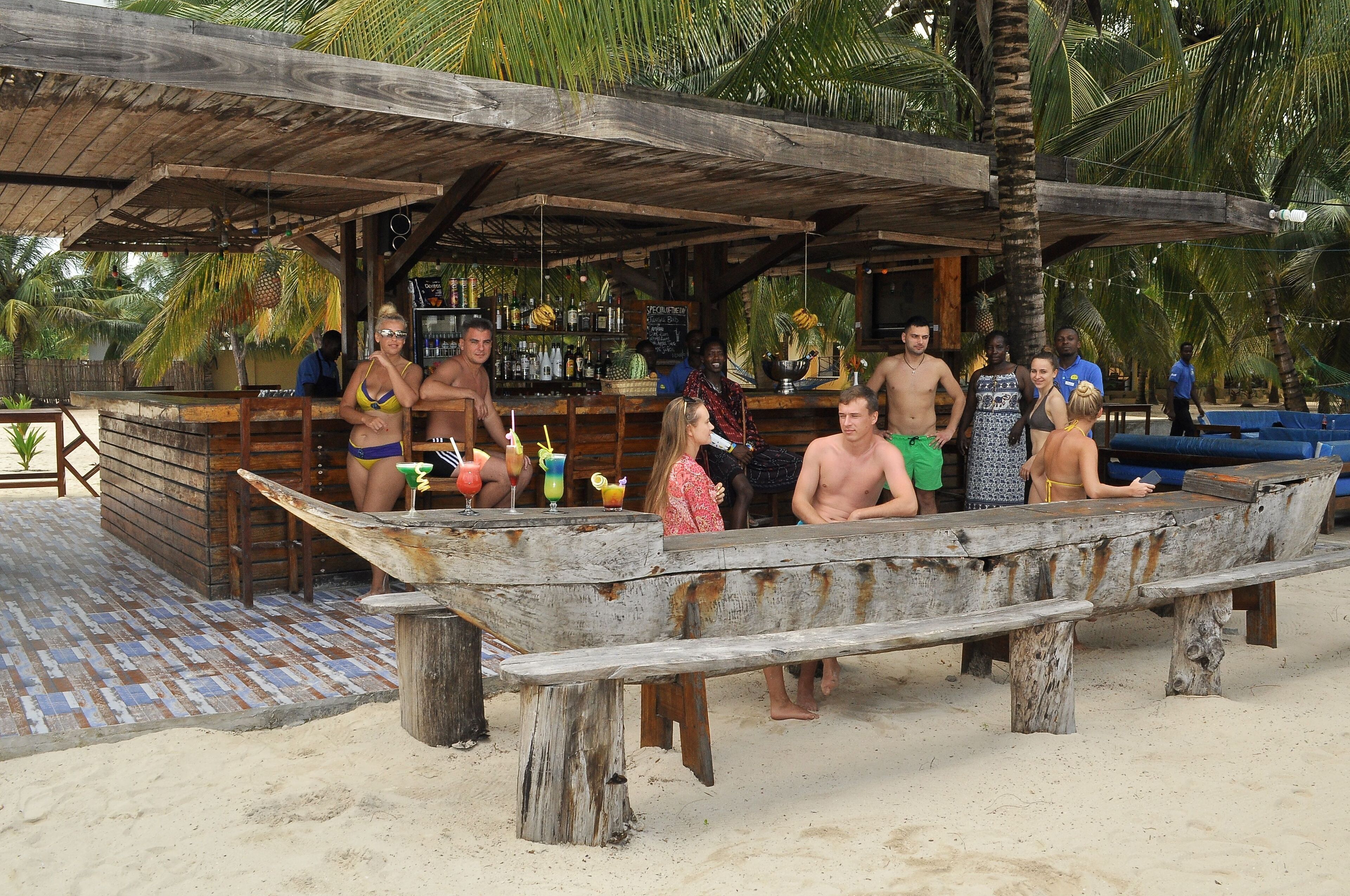 A wooden bar area with a rustic design, surrounded by palm trees and a sandy beach.