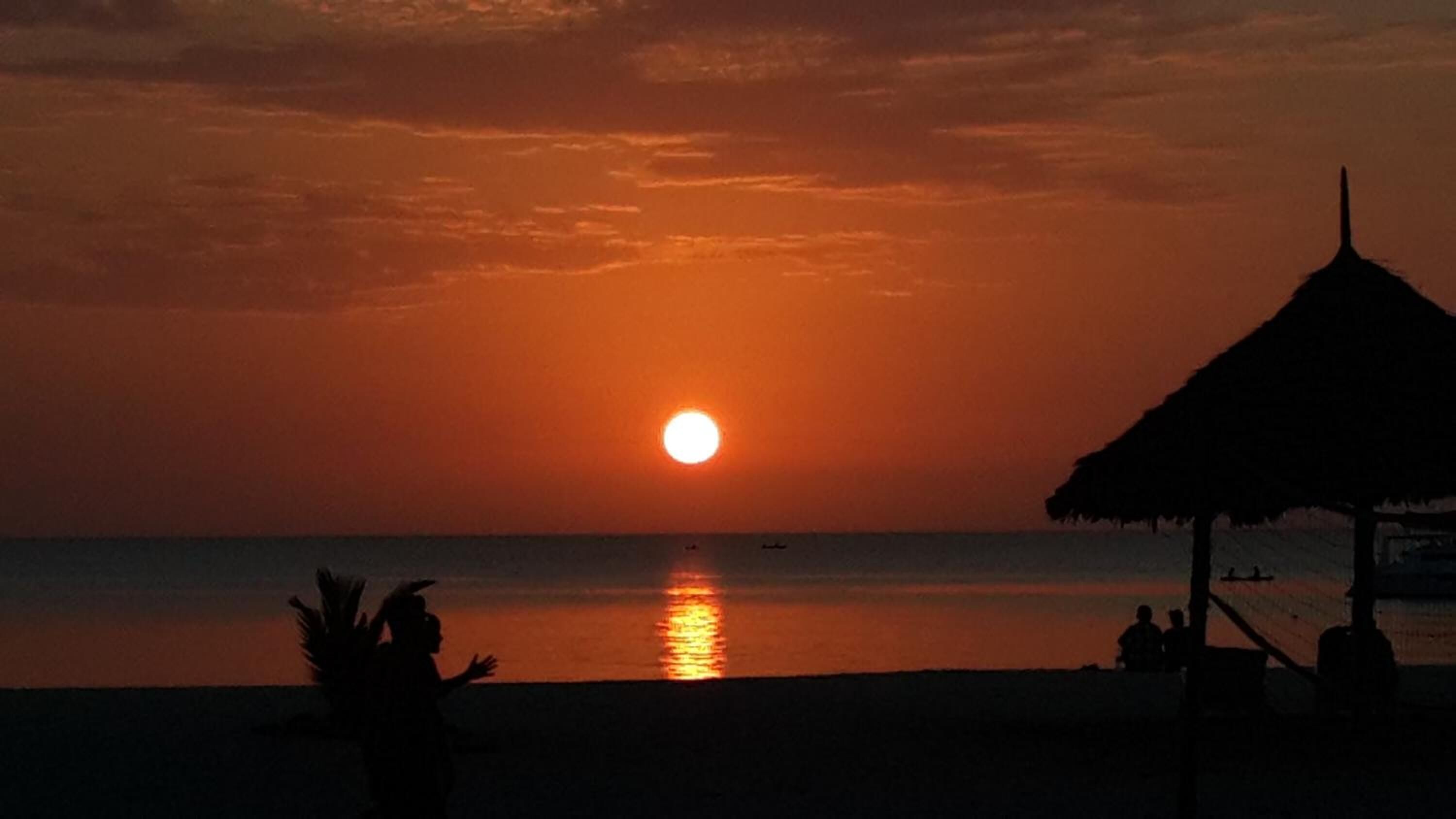 A sunset over a calm sea with a thatched-roof structure on the beach.