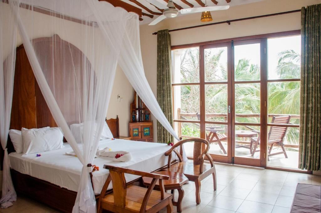 Una habitación con cama, una mesa y sillas, y un balcón con vistas a palmeras.