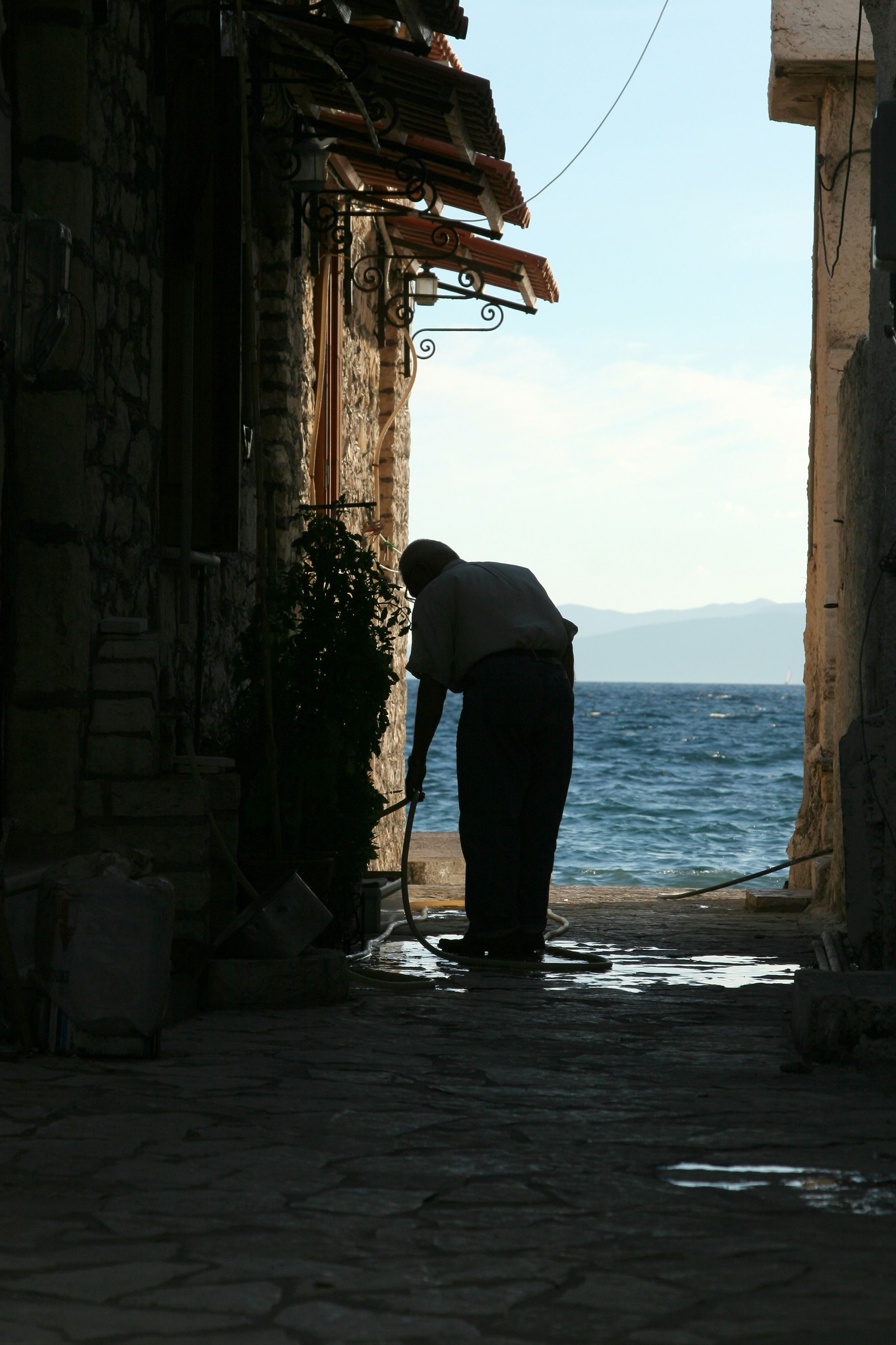 A narrow stone pathway leading to a body of water, with a person bending down, possibly cleaning or retrieving something.