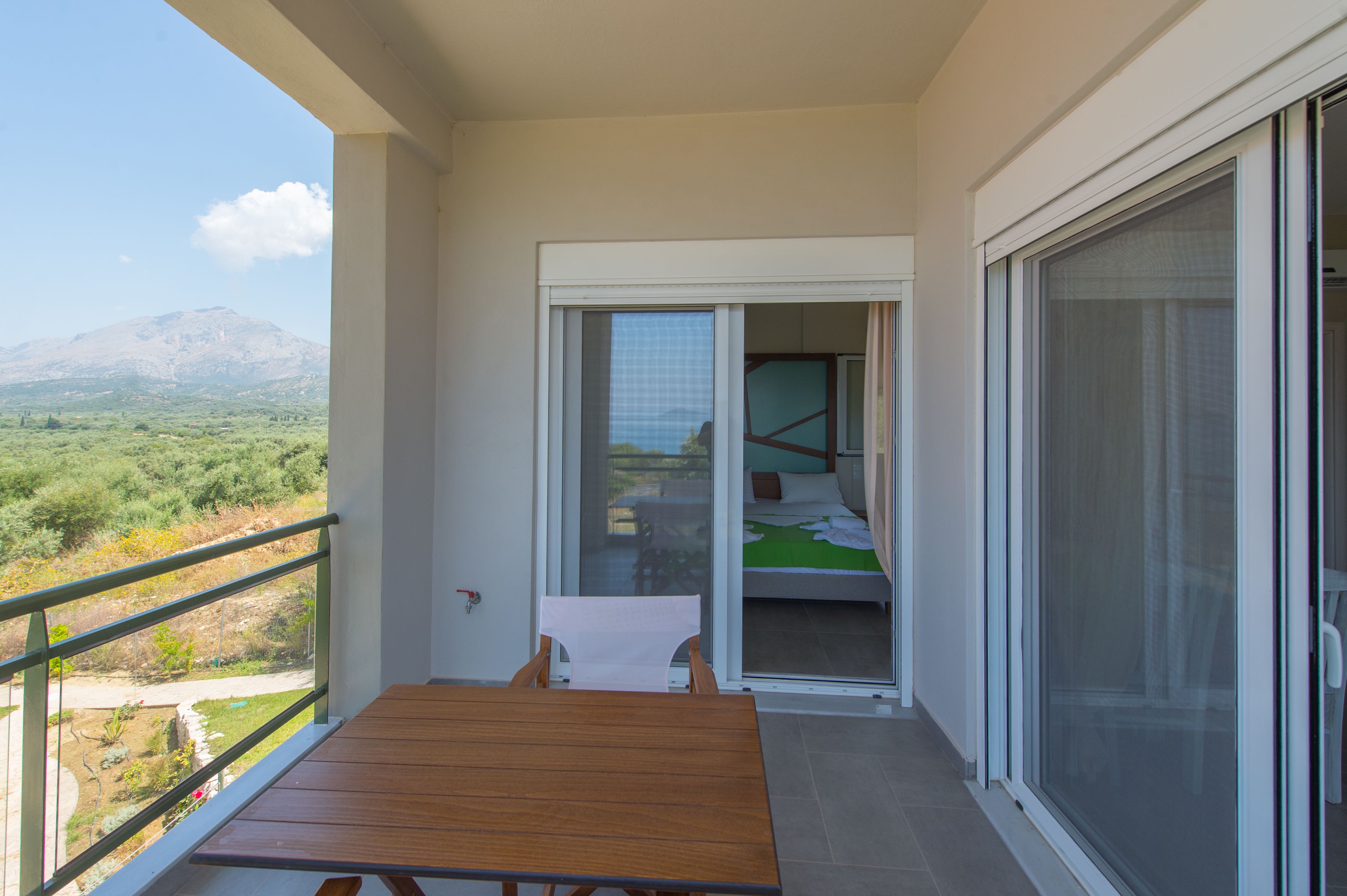 A balcony with a wooden table and chairs, a sliding glass door leading to a bedroom with a bed and green bedspread, and a view of a mountainous landscape.