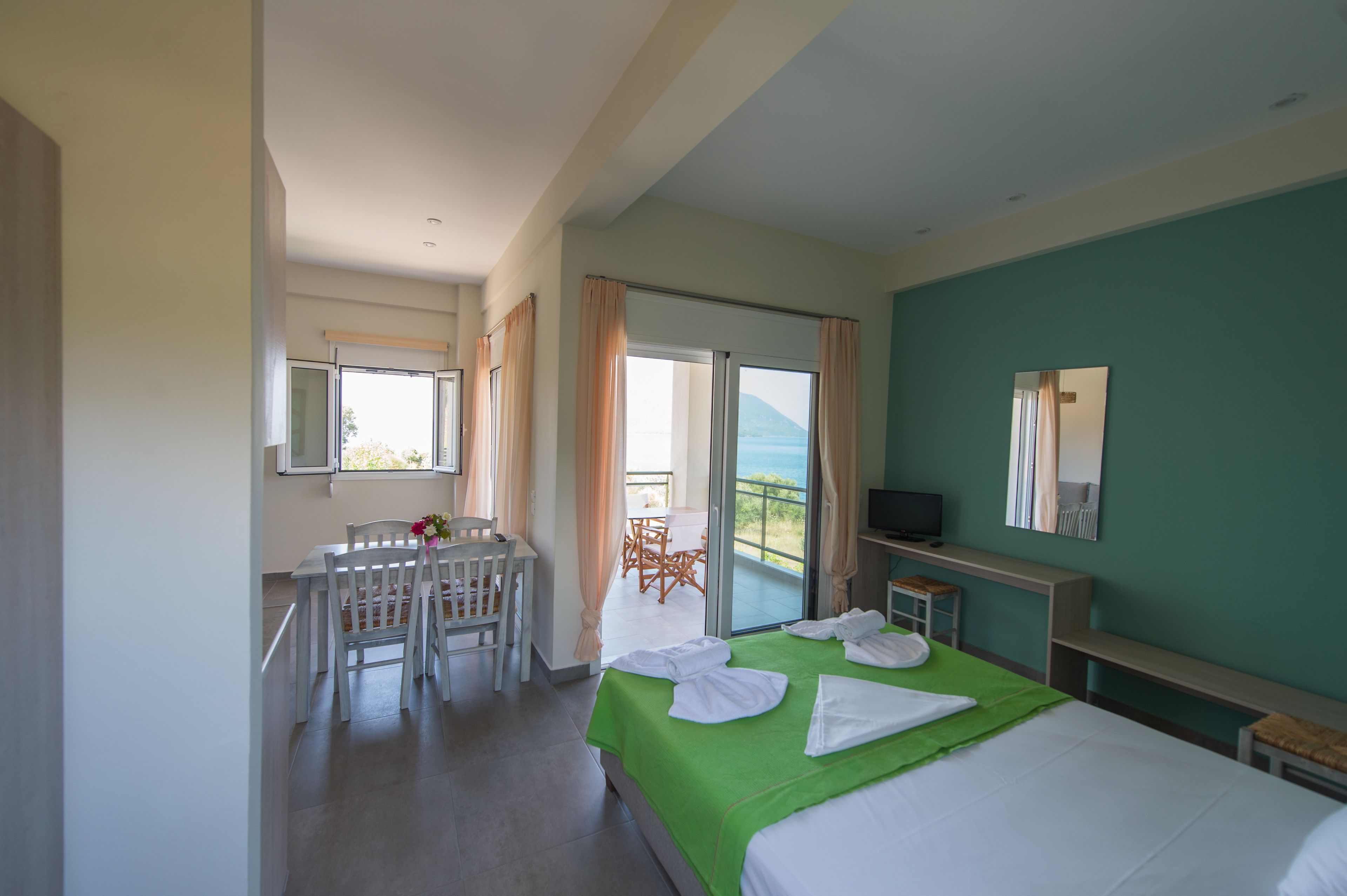A hotel room with a bed, a desk with a chair, a balcony with a table and chairs, and a television.