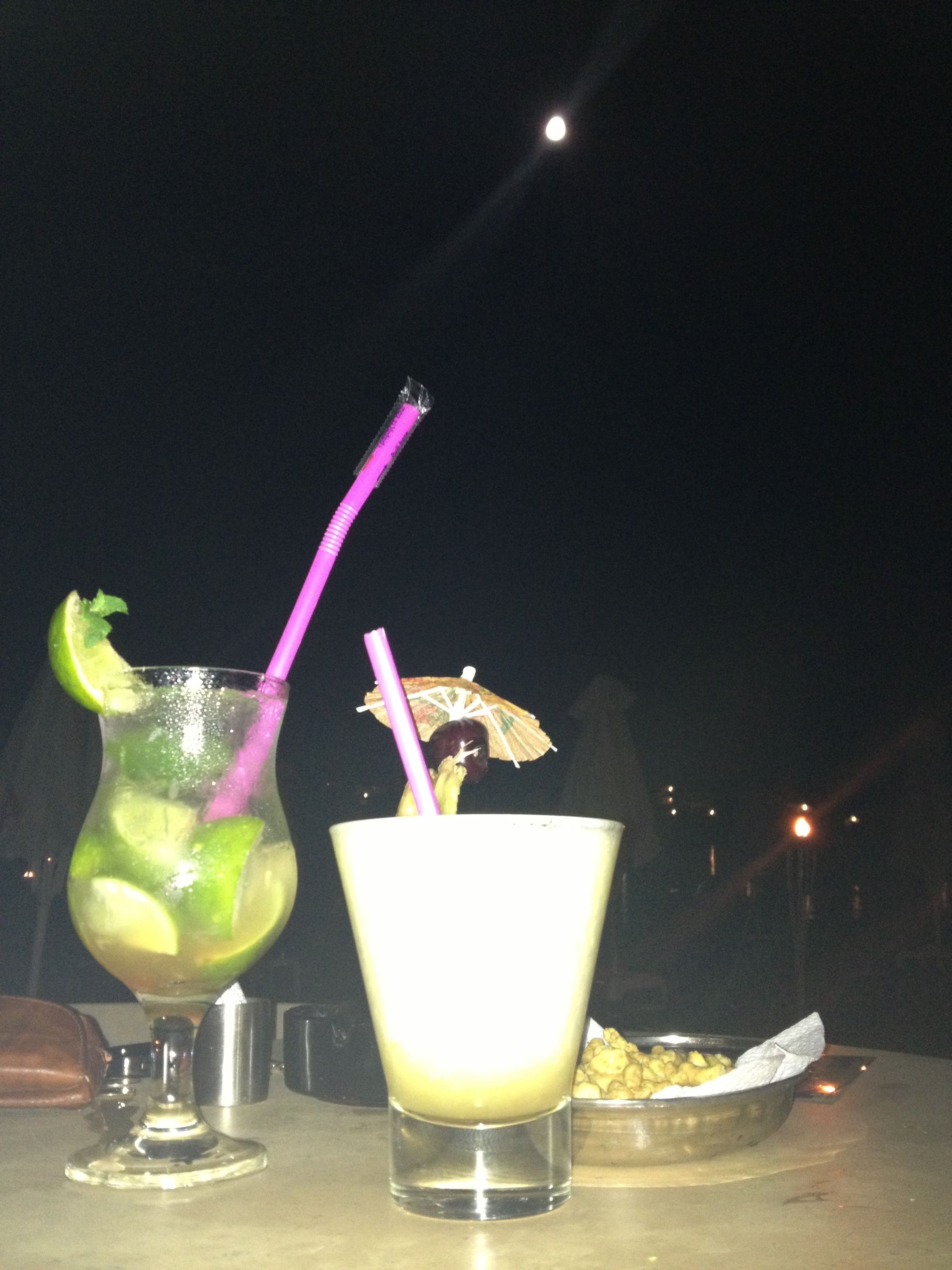 Dos bebidas sobre una mesa, con fondo oscuro y una luz tenue a lo lejos.