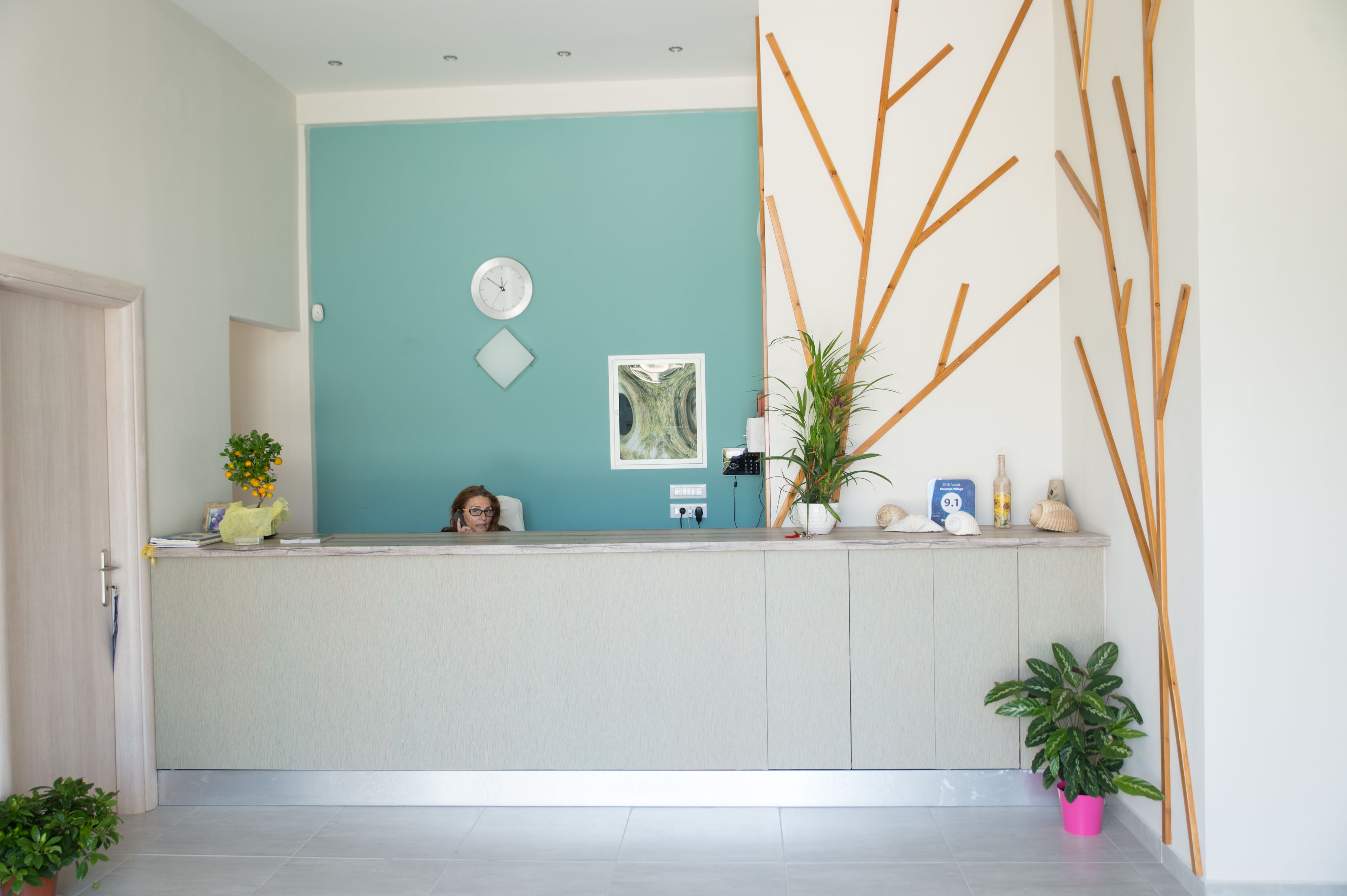 Un área de recepción con pared color verde azulado, un reloj, una obra de arte enmarco y una planta en maceta.