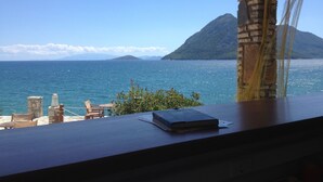 A view from a window overlooking a coastal scene with a mountain, a clear sky, and a table with a book and a chair. - Thesmos Village (Xiromero)