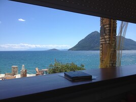 Vista desde una ventana sobre un paisaje costero con montaña, cielo despejado, una mesa, un libro y una silla.