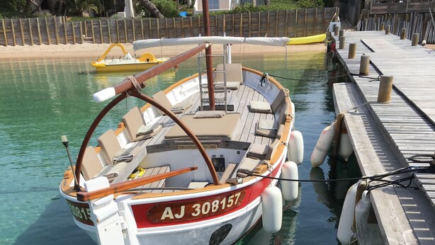 Un bote amarrado en un muelle, con agua cristalina, una cubierta de madera y una playa arenosa al fondo.