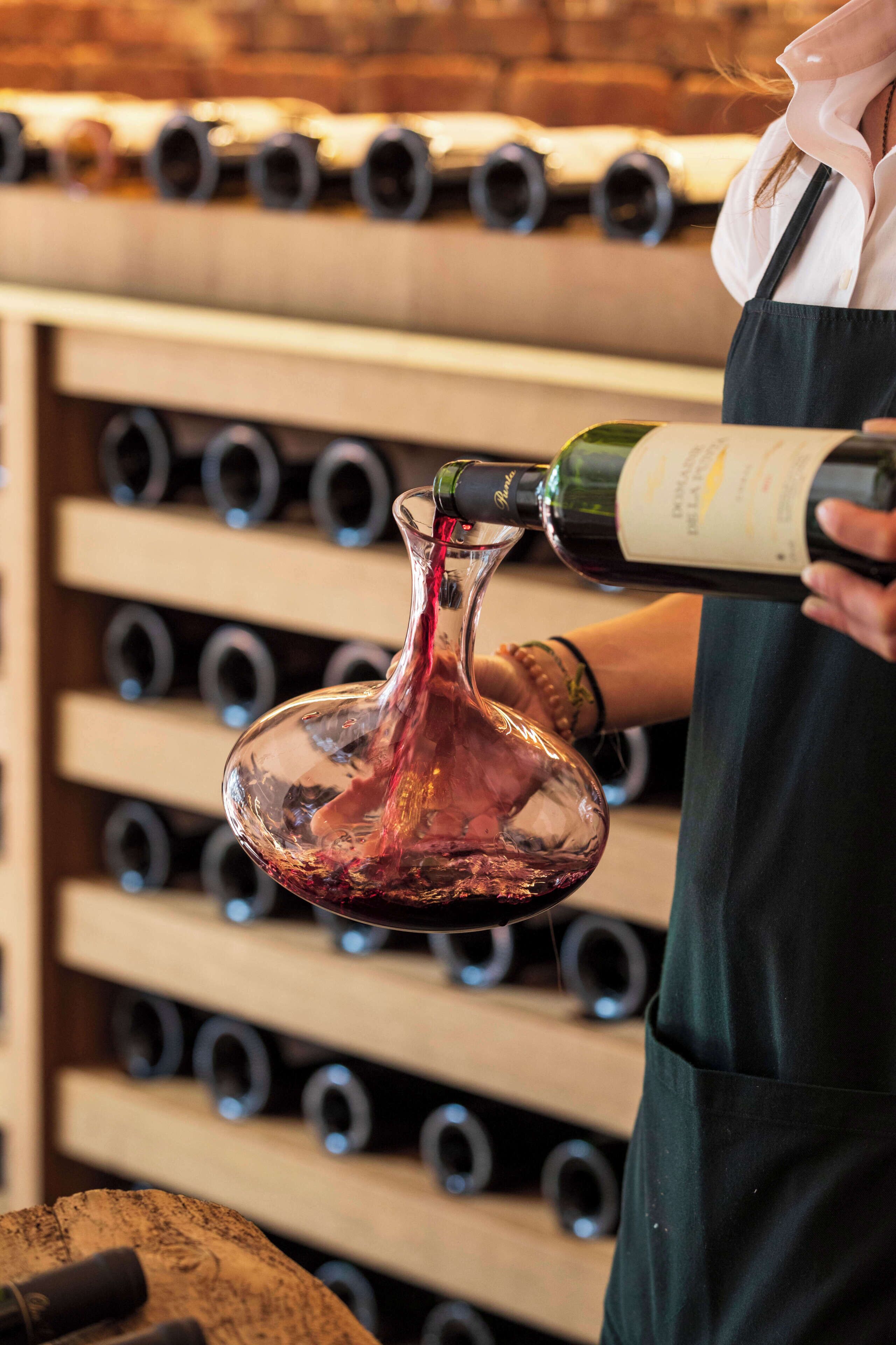 A person pouring wine into a glass, with a wine rack in the background.