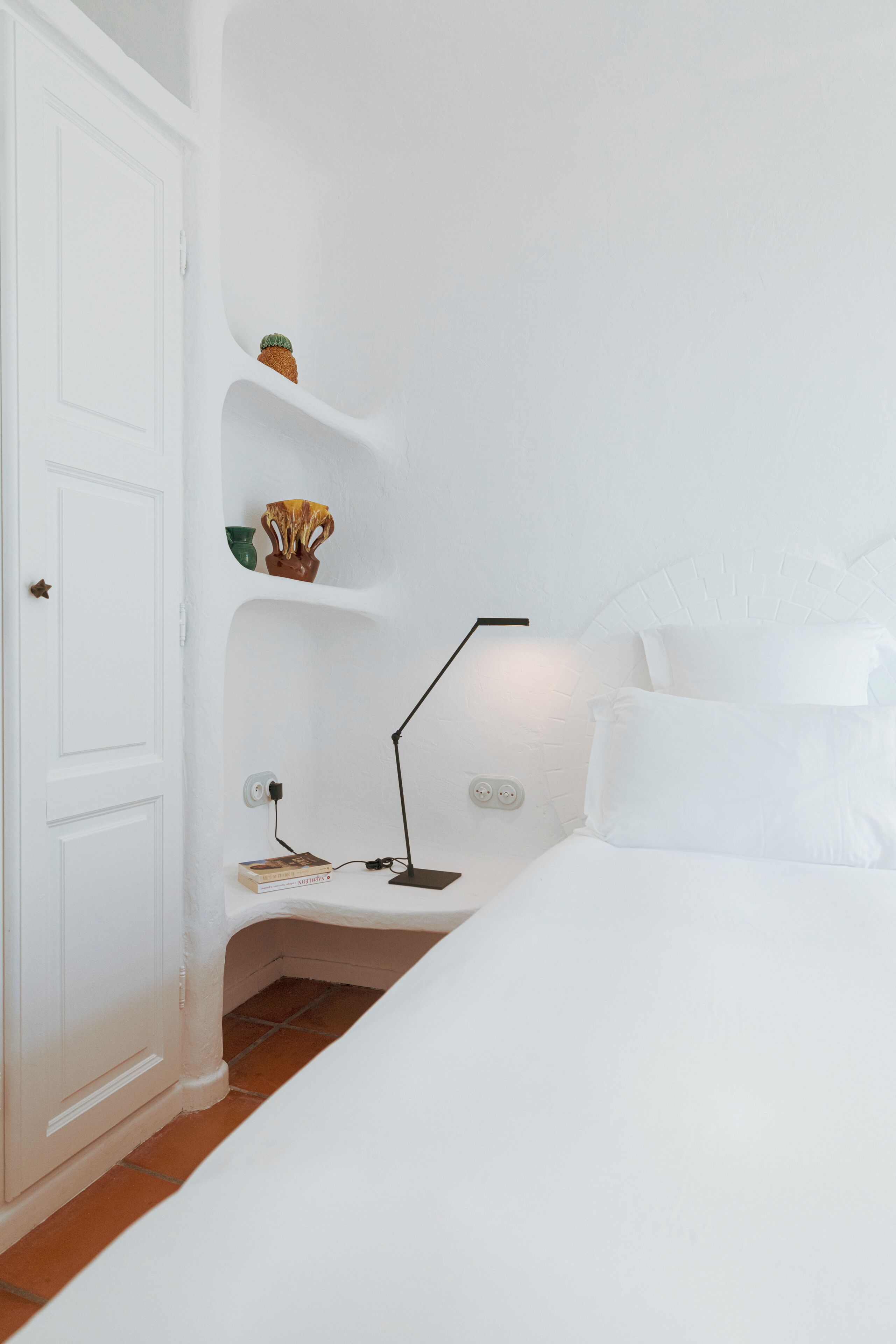 Une pièce blanche avec un lit, une table de chevet, une lampe et une porte.