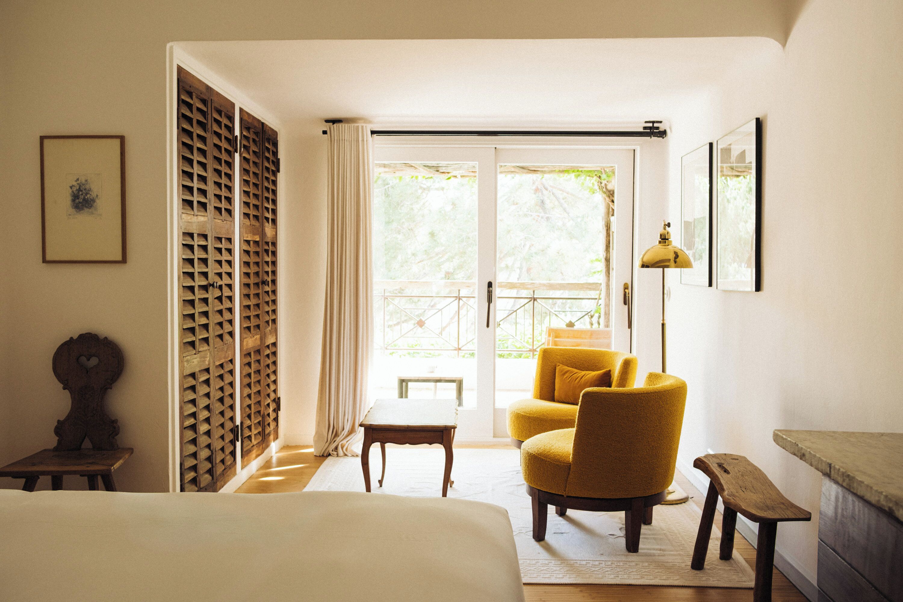 A room with a bed, a yellow armchair, a small table, a lamp, and a view through a glass door.