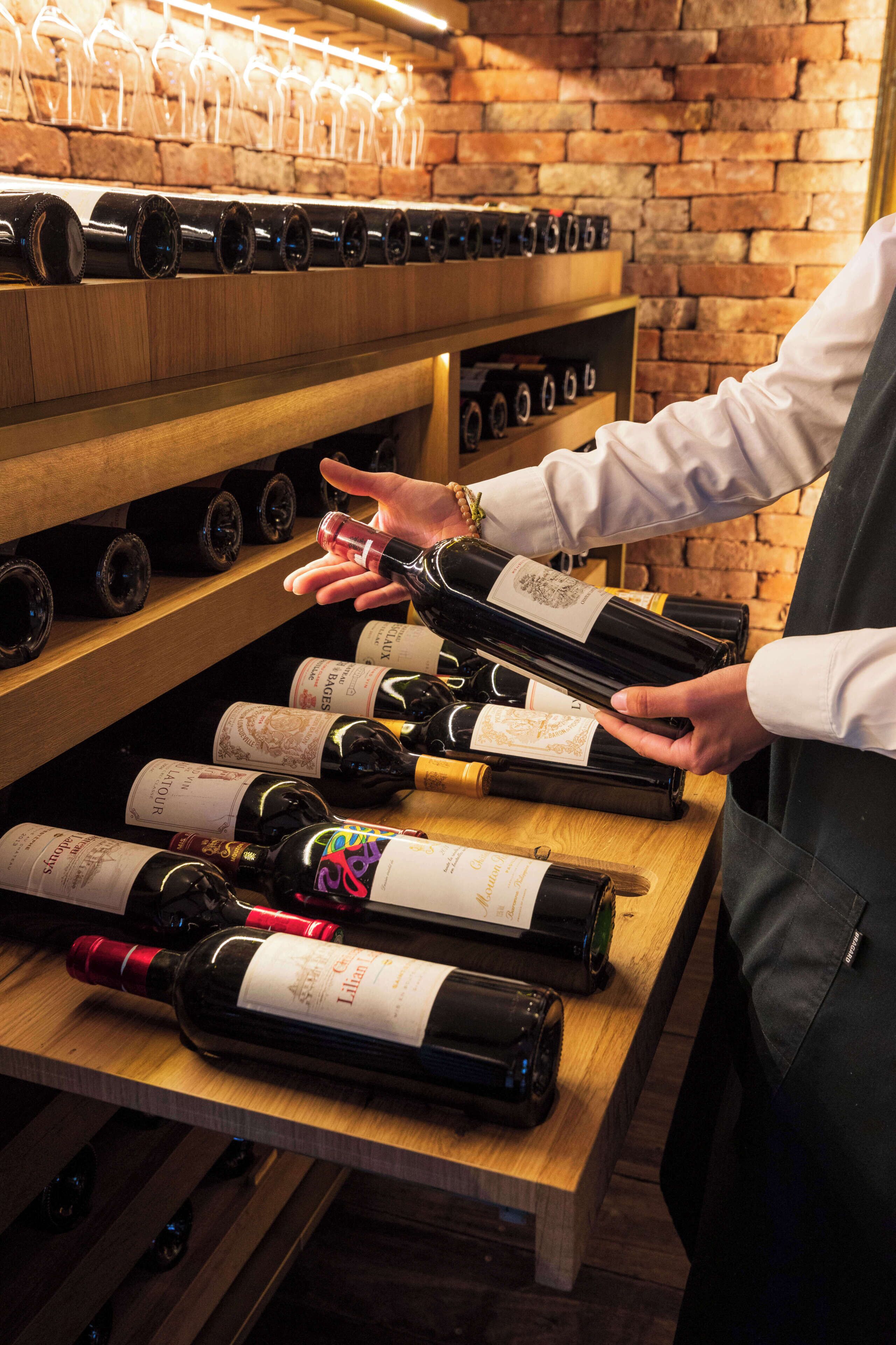 A person selecting a wine bottle from a wooden wine rack in a brick-walled room.