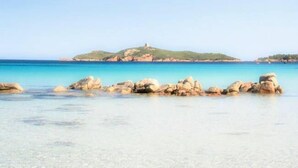 Una playa costera con agua turquesa cristalina, rocas grandes en primer plano y una colina con una estructura al fondo.