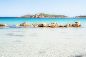 A coastal beach with clear turquoise water, large rocks in the foreground, and a hill with a structure in the background. - Grand Hôtel de Cala Rossa (Lecci)