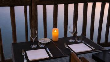 Mesa de comedor preparada para dos con una vela, copas y cubiertos, con vistas a una masa de agua tranquila donde se ven barcos y un muelle al fondo.