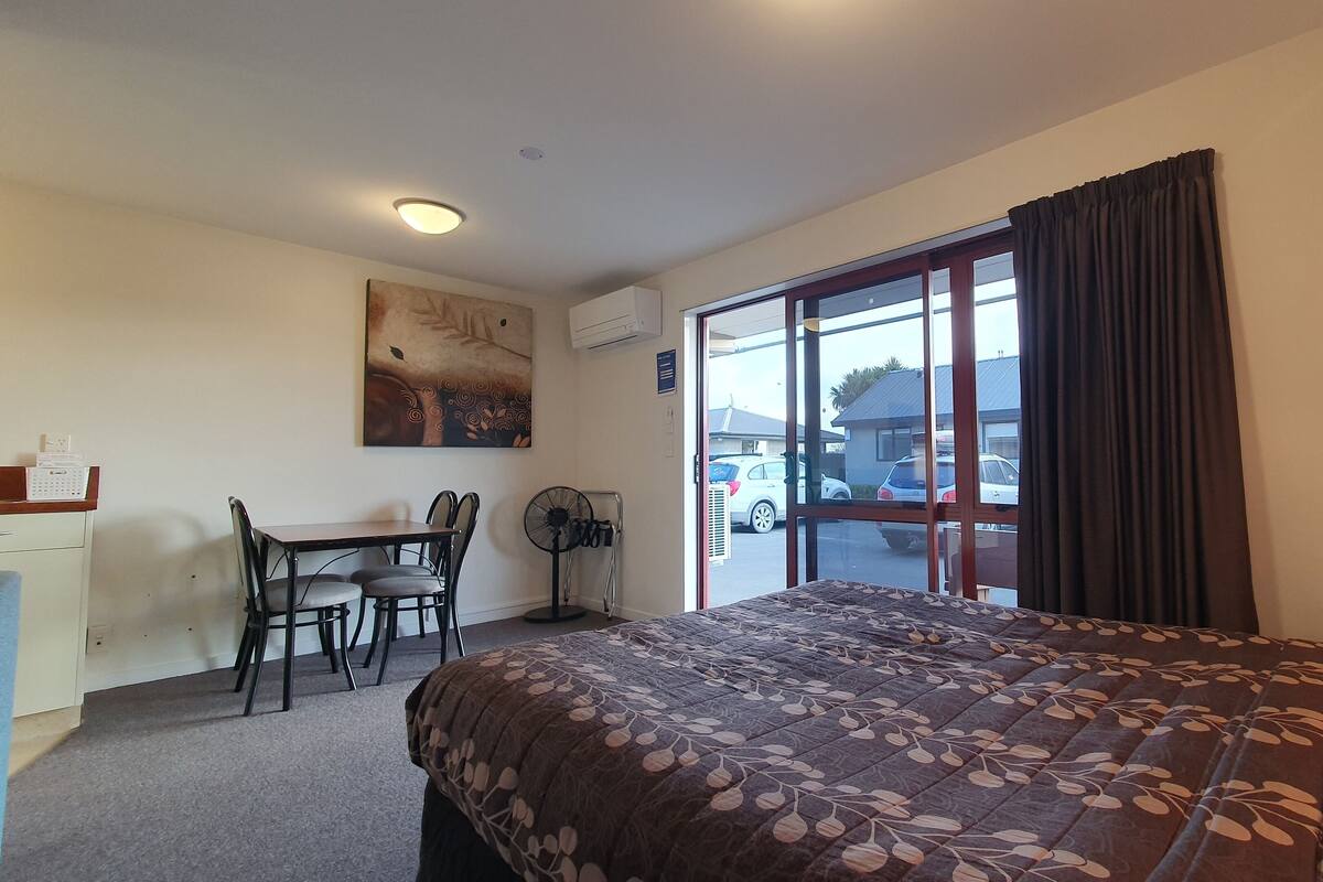 A hotel room with a bed, a desk with chairs, a standing fan, and a large window with curtains.
