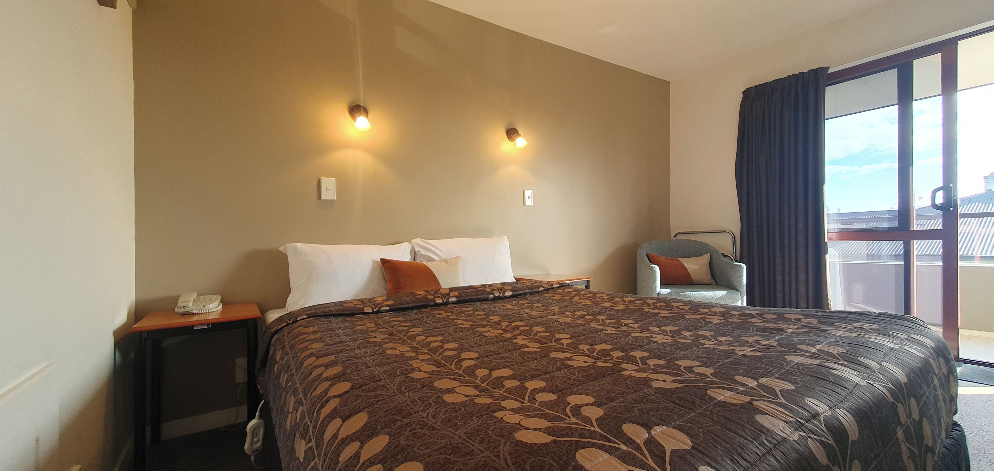 Habitación de hotel con una cama grande, dos lámparas de pared, una mesita con un teléfono y vistas al exterior a través de un ventanal.