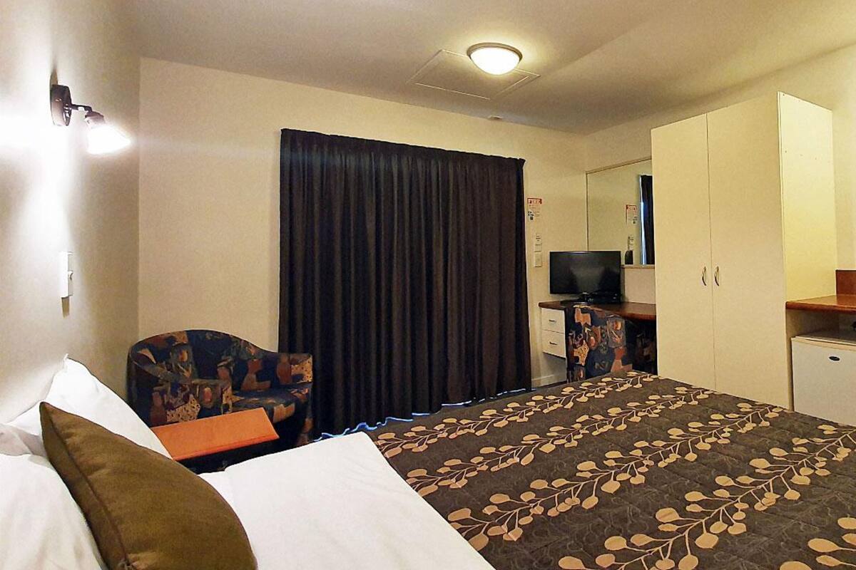 A hotel room with a bed, a desk with a television, a chair, a wardrobe, and a curtain.