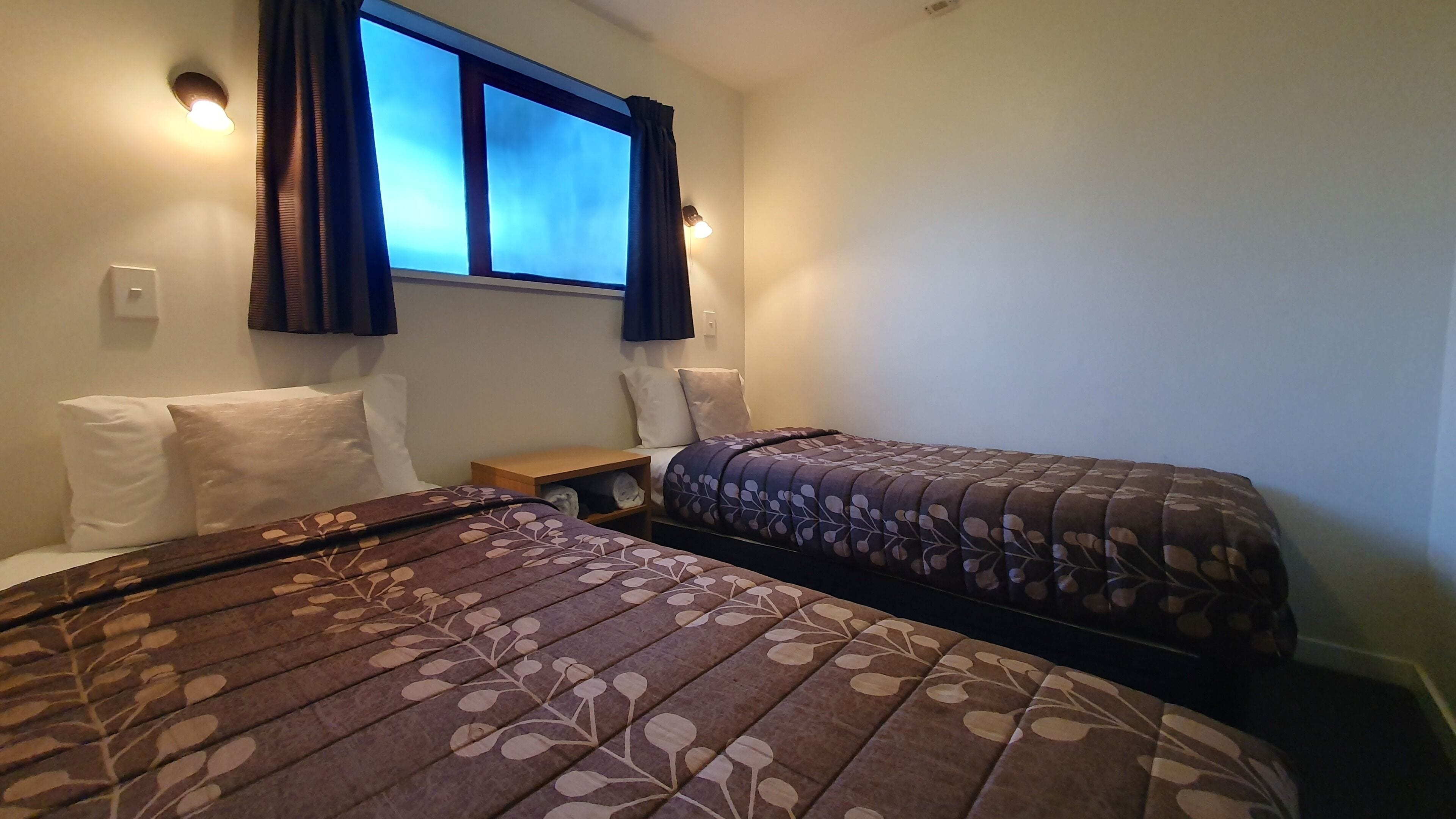 A hotel room with two beds, a window with curtains, a nightstand, and a wall-mounted light fixture.