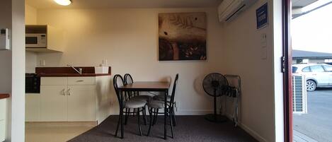 A compact kitchen with a microwave, sink, and a dining area with a table and chairs. There is a painting on the wall and a standing fan near the door.