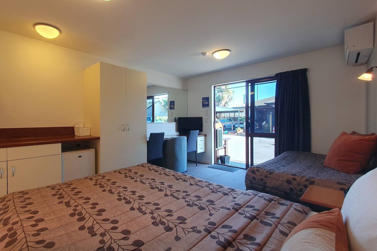 A hotel room with a kitchenette, a bed with a floral bedspread, a seating area with a table and chairs, and a view through a glass door.