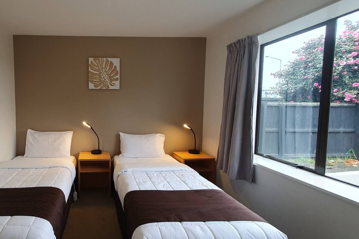 A hotel room with two single beds, a nightstand with a lamp, a wall-mounted artwork, and a window with a view of a garden.