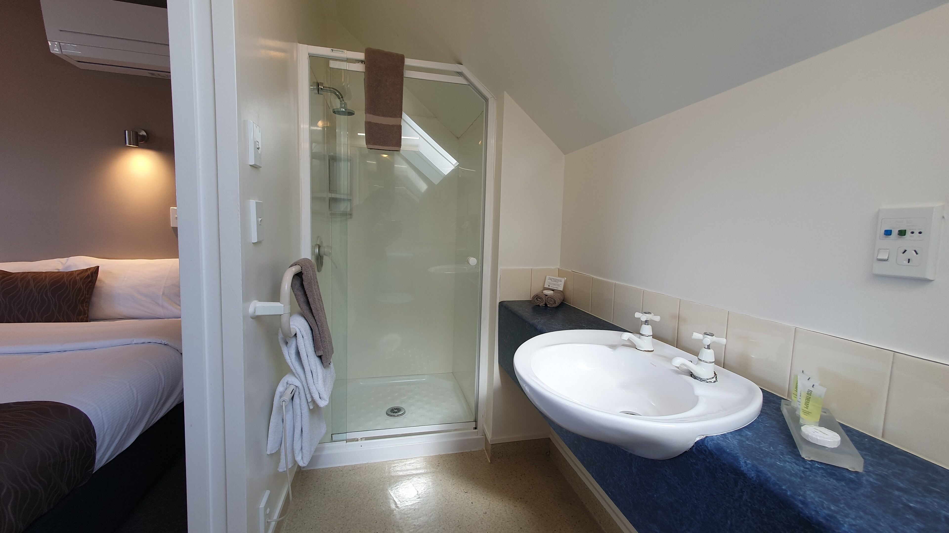 A modern bathroom with a glass-enclosed shower, a white sink, and a bed with a brown pillow.