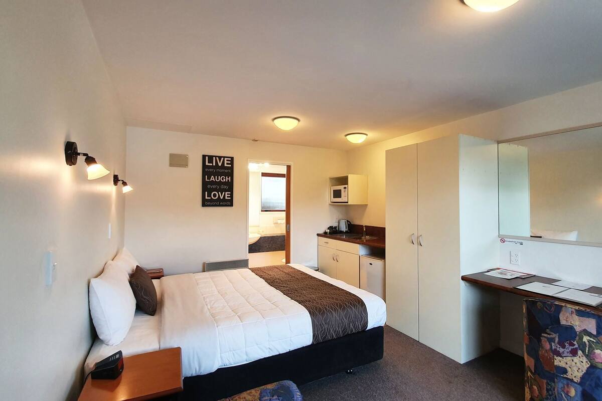 A hotel room with a bed, bedside table, a desk with a chair, a microwave, a cabinet, and a mirror.