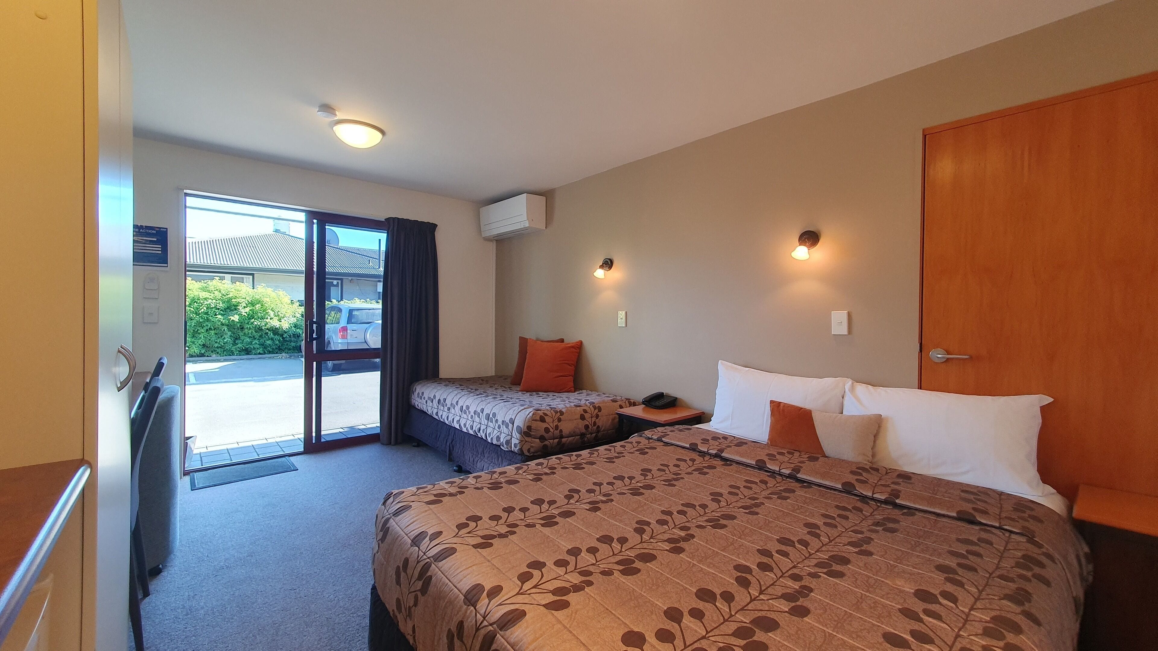 A hotel room with a large bed, a smaller bed, a desk, and a sliding glass door leading to a parking area.