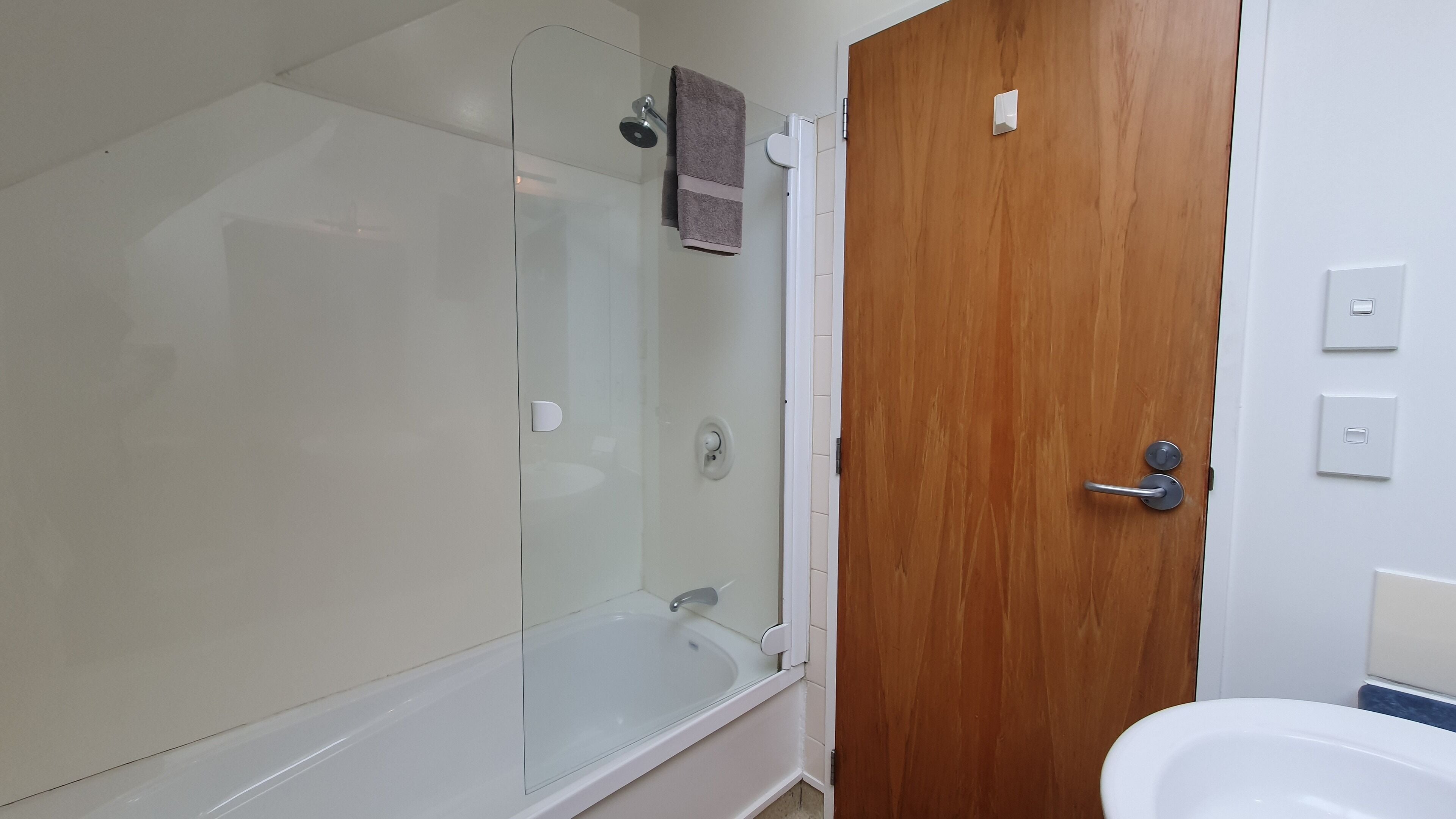 A bathroom with a glass shower enclosure, a wooden door, a white sink, and a towel hanging on the shower door.
