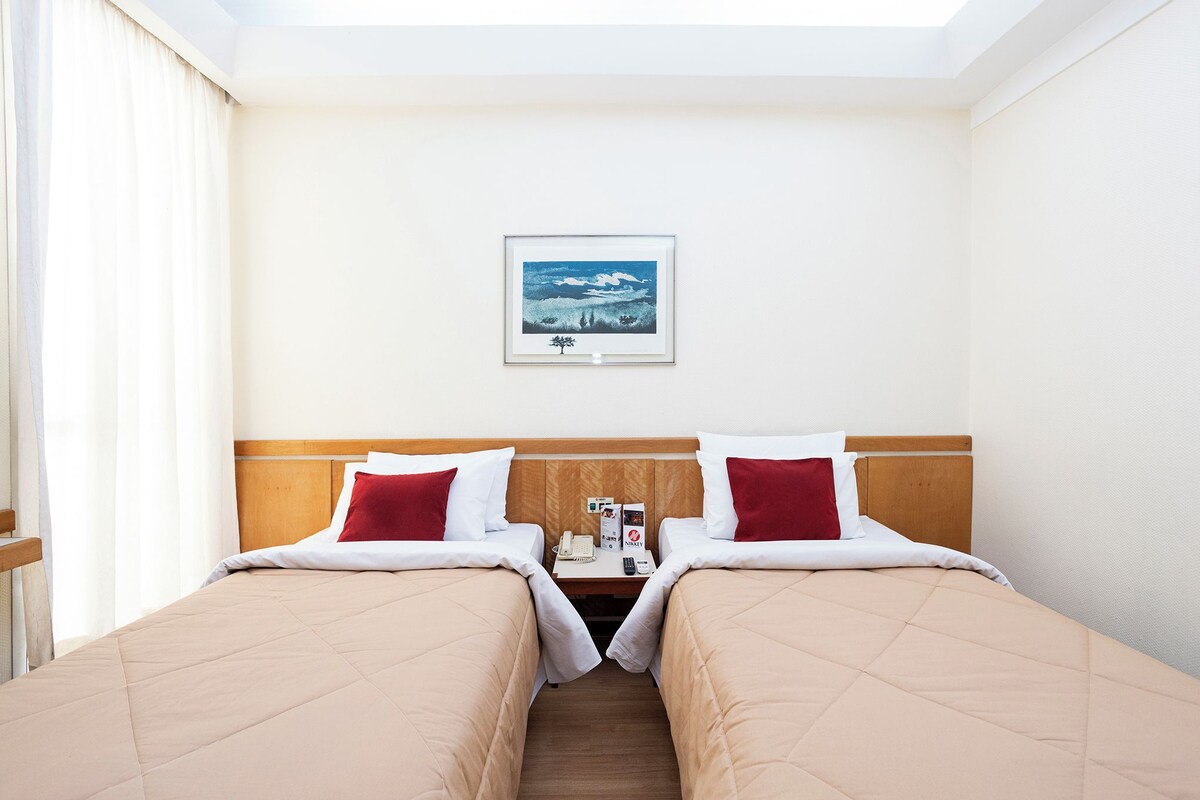 A hotel room with two beds, a wooden headboard, a nightstand with a phone and a box, and a framed picture on the wall.
