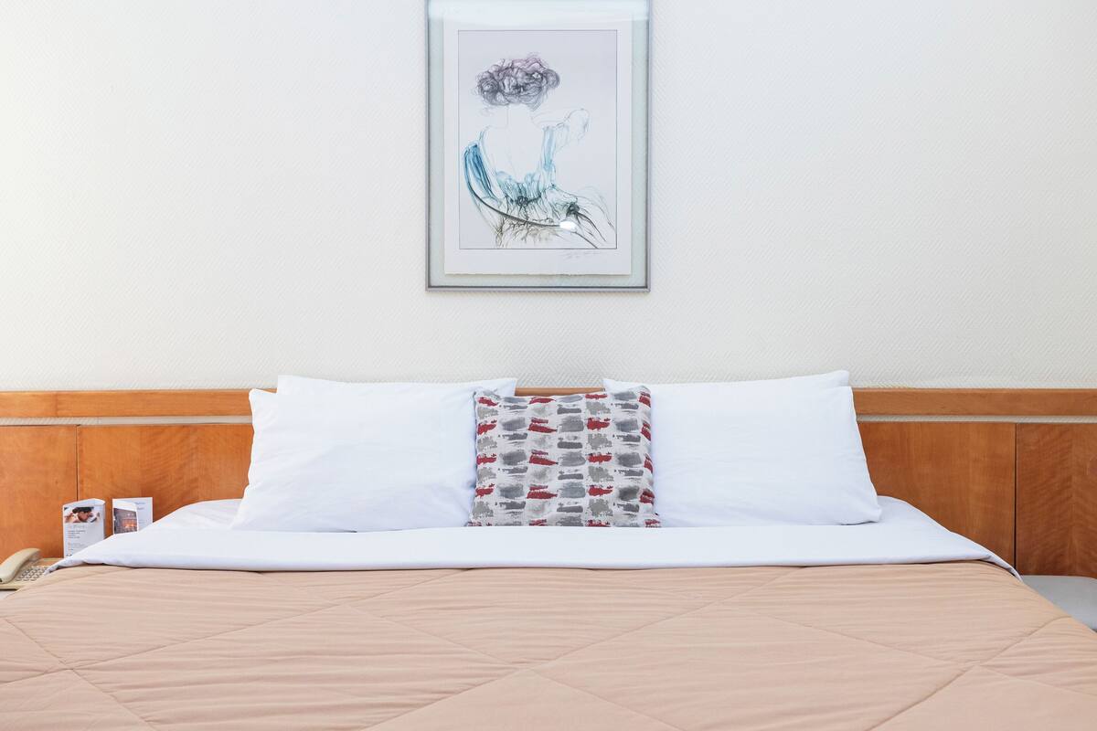 Ein ordentlich bezogenes Bett mit weißen und gemusterten Kissen, ein gerahmtes Kunstwerk darüber und ein Holz-Kopfstück.